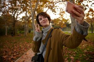 Positive young lovely short haired brunette woman raising hand with peace sign while making photo of herself with mobile phone, posing over yellowed trees in city garden