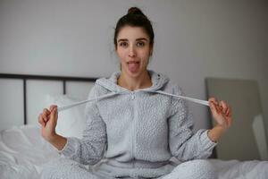 Indoor shot of lovely young dark haired woman with casual hairstyle looking joyfully to camera and showing tongue, sitting over home interior with crossed legs and pulling laces of her hoodie photo