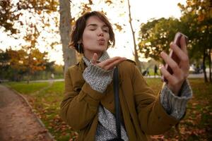 Outdoor shot of young pretty brown haired stylish woman with casual hairstyle raising her palm while blowing air kiss at camera of her mobile phone and winking with one eye photo