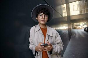 Portrait of young dark haired attractive female with nose piercing standing over city environment, holding mobile phone and looking ahead dreamily, wearing beige coat, foxy sweater and wide grey hat photo