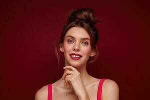 Studio shot of charming young brown haired female with bun hairstyle wearing festive makeup while posing over burgundy background, looking positively at camera and touching face gently photo