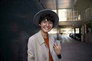 Beautiful dark haired stylish curly female looking at camera with charming smile, leaning on black city wall and keeping smartphone in hand, dressed in trendy wear and wide grey hat photo