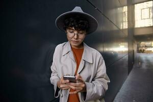 Photo of young dark haired stylish hipster female walking in city against black urban wall, keeping mobile phone and looking at screen with calm face, checking her maibox