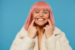 Portrait of young happy attractive pink haired lady with colored makeup keeping forefingers in corners of her mouth while smiling widely at camera, wearing white faux fur coat over blue background photo