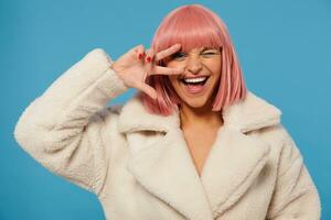 Studio photo of fancy cheerful young lovely lady with short pink haircut raising hand with victory sign to her face, keeping one eye closed while posing over blue background