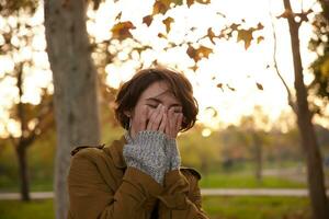 Joyful young pretty short haired brunette woman covering her face with raised palms while laughing happily, wearing stylish warm clothes while walking over yellowed trees in city garden photo