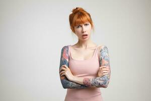 Bewildered young redhead tattooed female with bun hairstyle folding hands on her chest and frowning eyebrows while looking confusedly at camera, isolated over white background photo