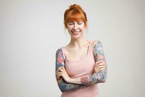 Pleasant looking young beautiful tattooed female being in nice mood and smiling happily with closed eyes, keeping hands crossed while posing over white background photo