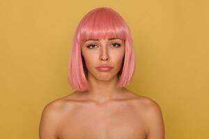 Wailful young attractive pink haired female with short haircut looking sadly at camera and pouting her lips while standing over mustard background with naked shoulders photo