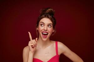 Attractive excited young brown haired female with evening makeup wearing her brown hair in bun and showing upwards positively with raised forefinger, posing over burgundy background photo