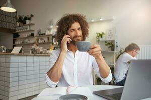 Young beautiful curly male with lush beard smiling cheerfully to camera and giving wink, drinking coffee during phone conversation, working in public place with laptop photo