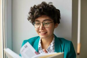 de cerca de positivo joven de ojos marrones corto peludo Rizado morena dama voltear emocionado libro y sonriente agradablemente mientras posando terminado blanco interior en soleado brillante día foto