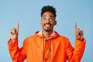 Young happy dark skinned attractive guy in orange rain coat looks up and wants to draw your attention pointing fingers up above his head, stands over blue background with copy space. photo