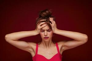 Displeased young brown haired female with bun hairstyle wearing pink top with straps while posing over burgundy background, frowning face and clutching her head, having headache after party photo