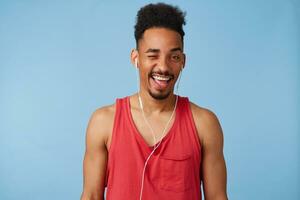 Young african american happy man looks at camera and winks, listens a new song of a popular group, wears in a red jersey, stands over blue background. photo