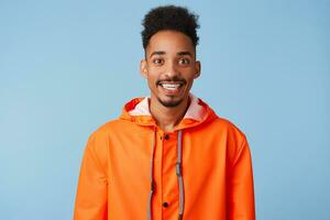 Close up of happy young attractive african american dark skinned boy wears in orange rain coat, smiles broadly stands over blue background. photo