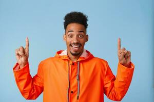 Hey, look up Happy young attractive african american man wears in orange rain coat, smiles broadly stands over blue background and wants to draw your attention at copy space, points fingers up. photo