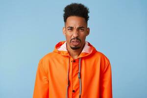 Close up of uhappy young african american dark skinned man in disgust, dissatisfied with the rainy weather outside, wears in orange rain coat, frowns isolated over blue background. photo