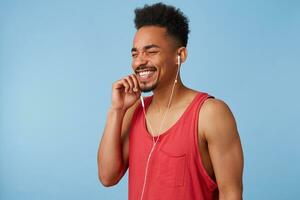 Portrait of young african american man music lovers feels good and very happy, close eyes, enjoys his favorite track list, sings along and dances stands over blue background. photo