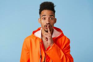 Close up of happy young african american dark skinned man in orange rain coat, tells secret information, demonstrates hush gesture, asks to keep quiet isolated over blue background. photo