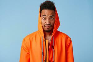 Outraged african american guy in orange rain coat narrows his eyebrows and clenches his lips, looks at the camera very displeased, stands over blue background. photo