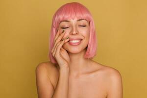 Pleased good looking young pink haired female with natural makeup touching gently her face with raised hand and smiling positively while posing over mustard background photo