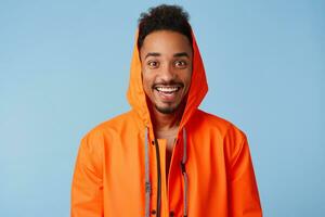 Close up of handsome african american dark skinned boy wears in orange rain coat, feels happy, smiles broadly and stands over blue background. photo