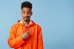 Unhappy young african american guy in orange rain coat looks at the camera inquiringly wants to draw your attention, points fingers at copy space on right. Stands over blue background. photo