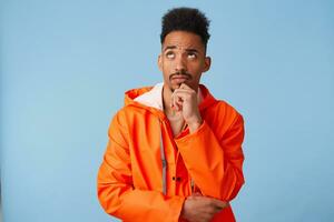 Portrait of young african american dark skinned man in orange rain coat, holds his chin, looks up and thinks about plans for the future stands over blue background. photo