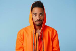 Sad handsome african american dark skinned boy wears in orange rain coat, confused, confused, lost train tickets and can not leave on time. Looks at the camera over blue background. photo