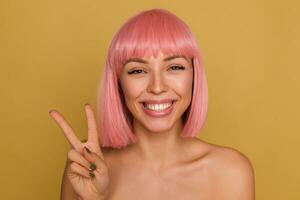 alegre joven atractivo dama con rosado de moda Beto peinado demostración su blanco Perfecto dientes mientras sonriente en general y demostración elevado dedos con victoria firmar, aislado terminado mostaza antecedentes foto