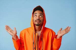 Portrait of young african american dark skinned guy in orange rain coat, hopes for good luck looking up raising, spreading his arms to the side, stands isolated over blue background with copy space. photo