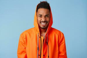 Frowning african american boy wears in orange rain coat grimacing, feel a terrible headache that is unbearable to endure, angry looking at camera standing over blue background. photo
