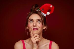 Close-up of charming blue-eyed young brunette lady with festive makeup wearing santa hat and looking dreamily upwards with pleasant smile, making new year wish over burgundy background photo