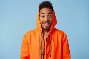 Photo of enthusiastic young african american dark skinned guy in orange rain coat, smiles broadly and looks into the camera with wide open mouth, stands over blue background.