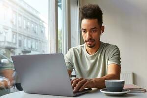 Photo of concentrate young attractive dark skinned boy, works at a laptop in a cafe, drinks coffee and thoughtfully looks at monitor.