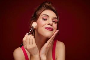 Close-up of pretty young brunette female with claret lips applying blushes on her cheeks while preparing for evening party with her friends, posing against burgundy background photo
