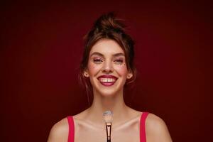 Close-up of positive attractive young brunette woman with bun hairstyle smiliing happily at camera and holding brush with lushes, making evening makeup while posing over burgundy background photo