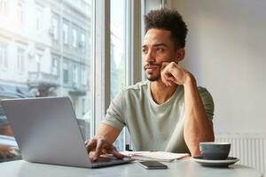 Portrait of young attractive African American thinking boy, sits at a table in a cafe, works at a laptop and drinks aromatic coffee and thoughtfully looks at the window. photo