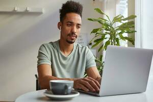 Young attractive African American pensive boy, sits at a table in a cafe, works at a laptop, looks at the monitor and trying to come up with a new topic for your blog. photo