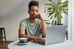 Portrait of frowning young attractive African American thinking boy, sits in a cafe, works at a laptop, touches cheek and sadly looks up, thinks about deadline. photo