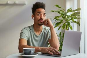 Portrait of young attractive African American thinking boy, sits in a cafe, works at a laptop and drinks aromatic coffee, touches cheek and dreamy looks up, thinks about the upcoming trip. photo