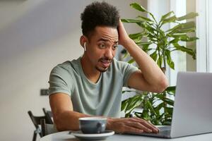 Young shocked attractive dark skinned boy, sits in a cafe and works at a laptop, looks at the monitor with surprised expression, read the article with dazed news. photo