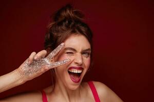 Joyful pretty young brunette female with claret lips wearing her hair in knot and raising victory sign with sparkles on fingers to her face, winking cheerfully at camera with wide mouth opened photo