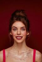 Portrait of beautiful young blue-eyed brunette lady wearing festive makeup and silver stars on her face while standing over burgundy background, looking positively at camera with sincere smile photo