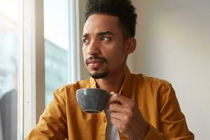 retrato de joven atractivo africano americano pensando chico, bebidas aromático café desde un gris leva y pensativamente mira lejos, molesto a gusto el gusto de grano. foto