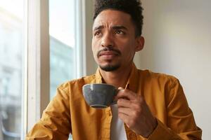 retrato de joven atractivo africano americano pensando chico, bebidas aromático café desde un gris taza , molesto a recuerda alguna cosa y pensativamente mira arriba. foto