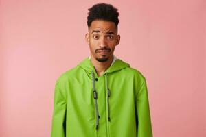 Displeased handsome dark skinned guy in green rain coat, raising his eyebrow questioningly, doubts and looks at the camera stands over pink background. photo
