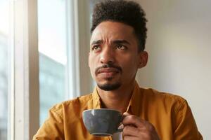 Portrait of young beauty African American thinking boy thoughtfully looks up, drinks aromatic coffee from a gray cup , trying to remember something photo