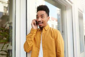 Young happy amazed dark skinned guy in yellow shirt walking down the street , speaks on the phone, hears unbelievable news, with wide open mouth and eyes, looks surprised. photo